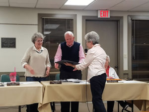 Three older adults stand behind a table at a meeting; one woman hands a framed item to a man while another woman watches and smiles. Another person is seated at the table. Nameplates and papers are visible.