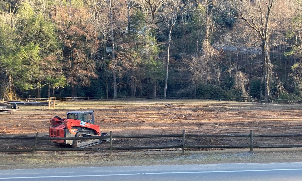 A red bulldozer sits on cleared, bare ground behind a wooden fence, with trees and brush lining the background and a road in the foreground.