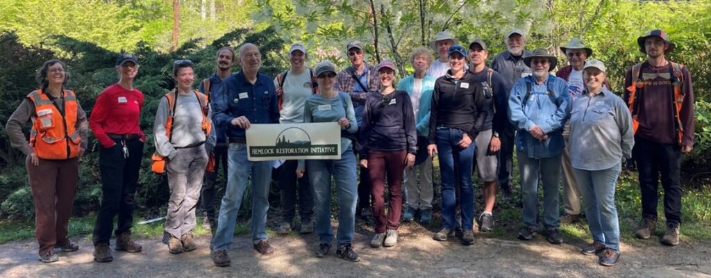 A group of about twenty people stand outdoors on a dirt path, posing and smiling for a photo. Some hold a sign that reads Wenlock Restoration Initiative. They are dressed in outdoor gear and surrounded by lush greenery.