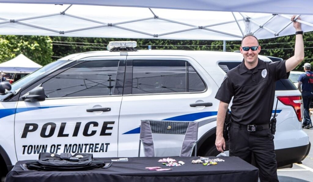 A police officer smiles under a canopy next to a Town of Montreat police SUV, standing behind a table with brochures and police gear at an outdoor event.