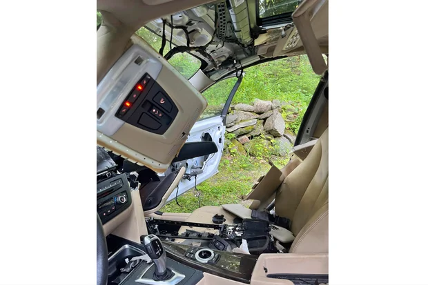 The interior of a heavily damaged car with exposed wires, missing panels, and detached ceiling controls. The driver’s door is open, and the seats and dashboard appear dismantled, with greenery visible outside.