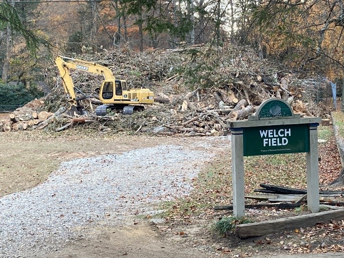 A yellow excavator sits atop a large pile of cut tree branches and logs, with a green Welch Field sign in the foreground. Trees with autumn foliage are visible in the background.