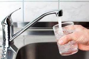 A hand holds a glass under a running kitchen faucet, filling it with water. The scene shows a stainless steel sink and tiled backsplash in the background.