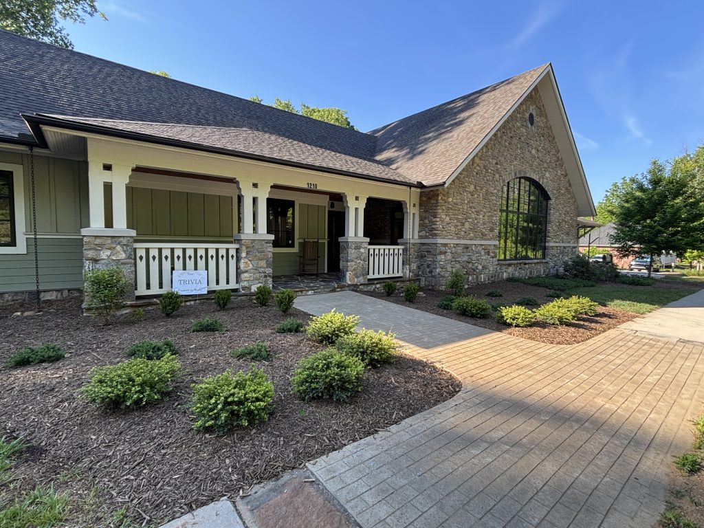 A single-story building with a stone façade, large arched window, and covered porch, surrounded by trimmed shrubs and mulch beds, under a clear blue sky. A sign reading TRIVIA is near the entrance.