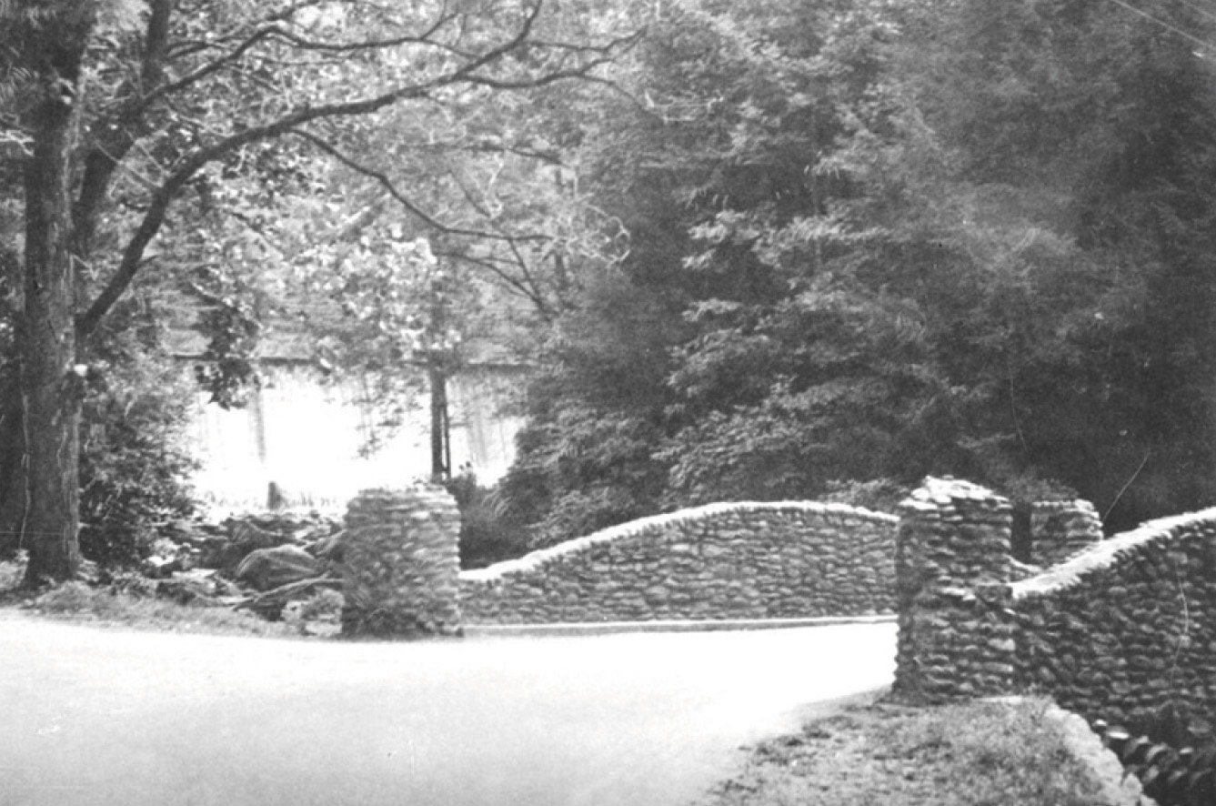 A black and white photo of a small, curved stone bridge surrounded by dense trees and foliage, with sunlight filtering through the branches and a quiet, winding road leading over the bridge.