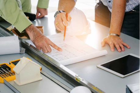 Two people review architectural blueprints on a desk with a tablet, measuring tape, a small model house, and a white hard hat nearby, suggesting a construction or design planning meeting.