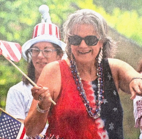 Two women celebrate outdoors; one wears sunglasses, patriotic beads, and a red, white, and blue dress, while waving an American flag. The other wears a stars-and-stripes hat and glasses. Both are smiling and festive.