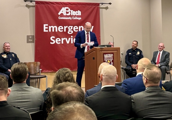 A man in a suit speaks at a podium in front of an audience at an AB Tech Community College event. Behind him is a red banner that reads Emergency Services. Several people, including police officers, are seated on stage.