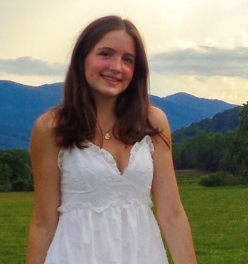 A young woman with long brown hair wearing a white dress stands smiling in a grassy field with mountains and a cloudy sky in the background.