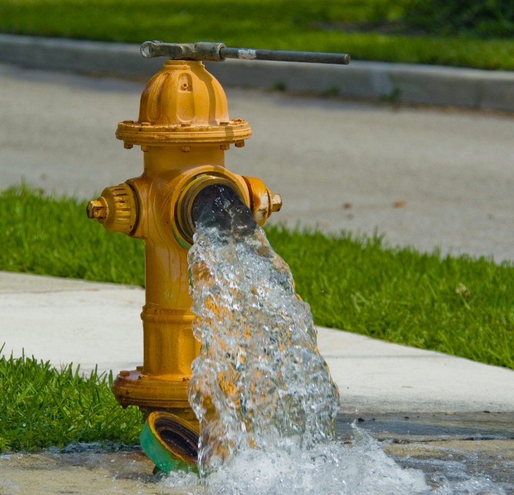 A yellow fire hydrant with its cap removed gushes water onto a sidewalk next to green grass, with a street and curb in the background.