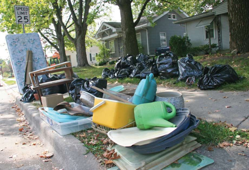 Household items such as furniture, bins, and a mattress are piled on a curb, along with many black garbage bags, in front of houses and beneath trees on a residential street with a 25 mph speed limit sign.