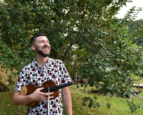 A man wearing a floral shirt stands outside, smiling and holding a violin in front of a leafy green tree.