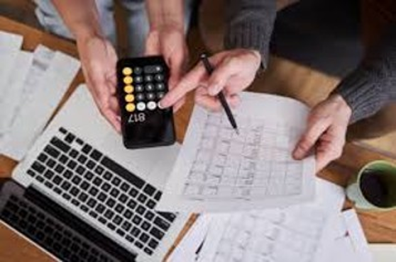 Two people work at a desk with a laptop, papers, a coffee cup, and a smartphone showing a calculator app with the number 817. One person points at a paper while the other holds the phone.