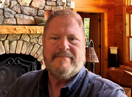 A middle-aged man with a beard and short hair smiles slightly, posing indoors in front of a stone fireplace and wood-paneled walls in a cozy, rustic room.