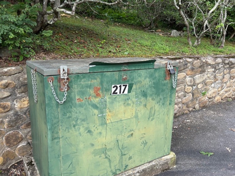 A weathered green metal utility box with chains and the number 217 sits in front of a stone wall and greenery.