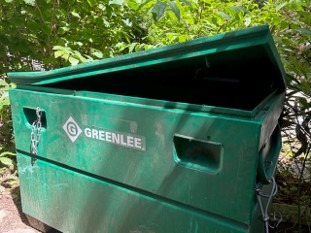 A green metal GREENLEE storage container sits outdoors with its lid partially open, surrounded by leafy plants.
