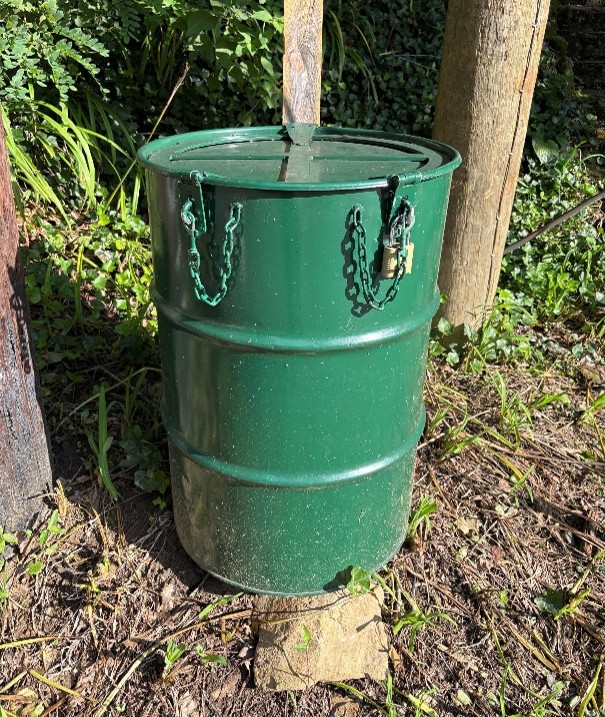 A green metal barrel with a lid secured by chains and a padlock stands on a stone outdoors, surrounded by grass and wooden posts in a shaded area.