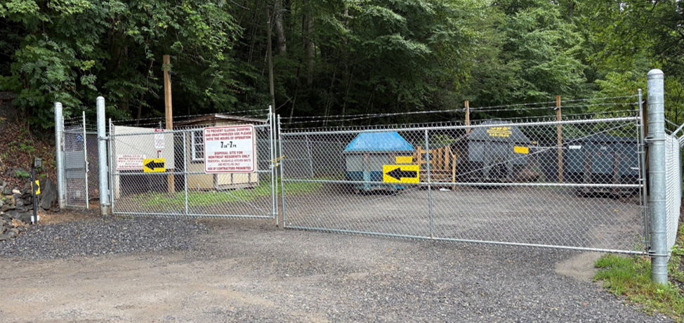 A gated area with dumpsters and recycling bins behind a chain-link fence, surrounded by trees. Signs with arrows and information are posted on the fence and buildings. The ground is gravel.
