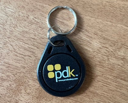 A black key fob with a metal keyring on a wooden surface. The fob has a yellow pdk logo and website text displayed on its front.