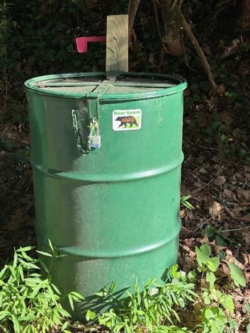 A green metal barrel with a locked lid sits outdoors. A sticker showing a bear and the words Bear Aware is on the barrel. There is foliage and sunlight on the ground around it.