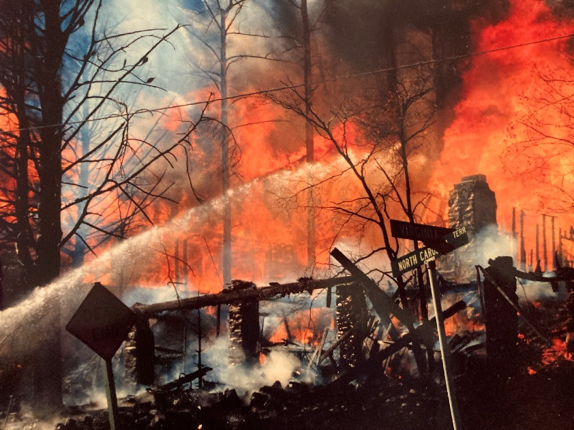 Firefighters spray water on a raging wildfire engulfing trees and ruins of buildings. Street signs marked North Carolina and Tennessee are visible in the foreground, with thick smoke and flames dominating the background.