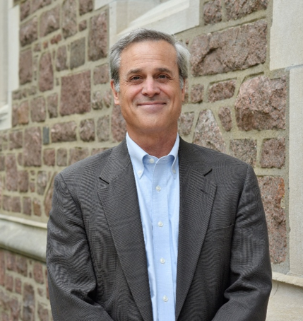 A man in a light blue button-down shirt and dark blazer standing in front of a stone wall.