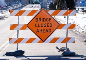 A roadblock with orange and white stripes and a sign that reads Bridge Closed Ahead blocks a snow-lined street.