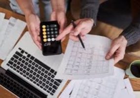 Two people work at a desk with a laptop, papers, and a cup. One holds a phone displaying a calculator app, while the other points at a document with a pen, suggesting financial or budget planning.
