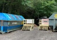 Two large dumpsters—one blue and one gray—sit on a gravel area with wooden platforms and signs in front of them, surrounded by dense green trees and a small building in the background.