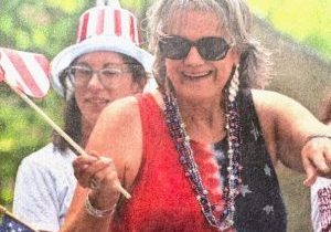 Two women celebrate outdoors; one wears sunglasses, patriotic beads, and a red, white, and blue dress, while waving an American flag. The other wears a stars-and-stripes hat and glasses. Both are smiling and festive.
