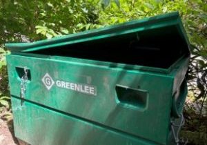 A green metal GREENLEE storage container sits outdoors with its lid partially open, surrounded by leafy plants.