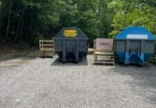 Three large dumpsters—one black, one brown, and one blue—sit on a gravel lot surrounded by trees. Wooden pallets are placed on the ground in front of the dumpsters. The sky is partly cloudy.
