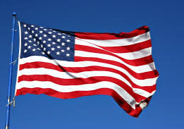 The flag of the United States, featuring red and white stripes and a blue field with white stars, waves in the wind against a clear blue sky.