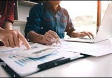Two people work at a desk with a laptop and financial documents. One points to a chart with graphs, while the other uses the laptop, suggesting a collaborative business or financial discussion.