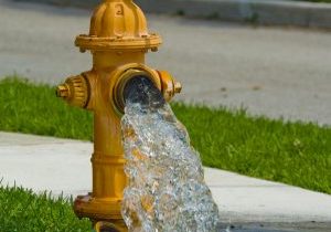 A yellow fire hydrant with its cap removed gushes water onto a sidewalk next to green grass, with a street and curb in the background.