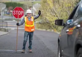 A construction worker in a safety vest and hard hat holds a stop sign and raises his hand, signaling a black car to stop on a road. Trees and a building appear in the background.