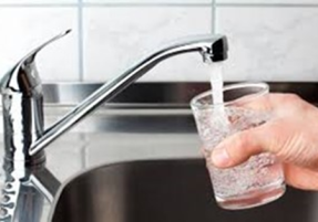 A hand holds a glass under a running kitchen faucet, filling it with water. The scene shows a stainless steel sink and tiled backsplash in the background.