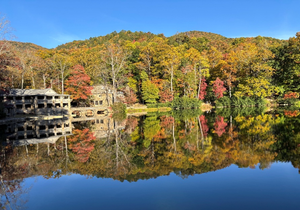 A stone building sits on the shore of a lake, reflecting the vibrant autumn colors of the surrounding trees in the calm water.