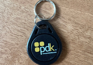 A black key fob with a metal key ring, featuring the yellow pdk logo and the website www.prodatakey.com, placed on a wooden surface.